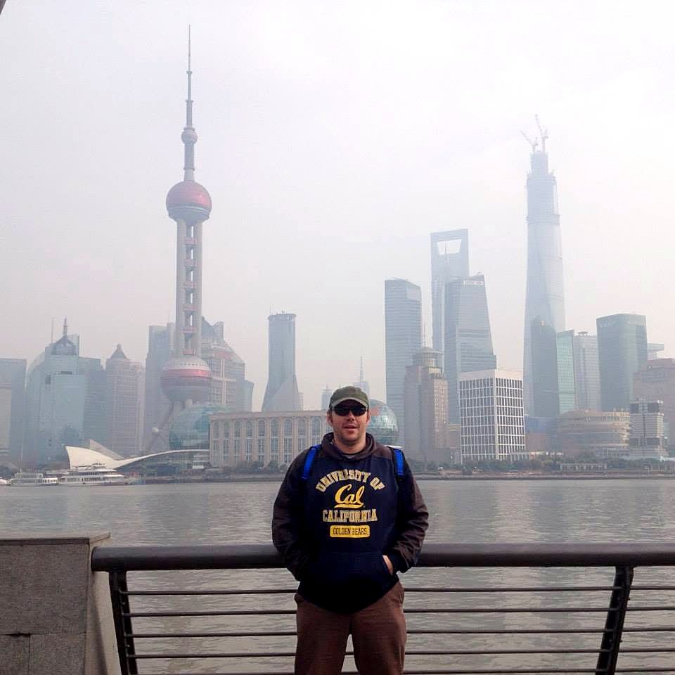 Ed standing on the Bund in Shanghai in a cap and Cal hoodie, with the Pudong skyline and Oriental Pearl Tower behind him across the river.